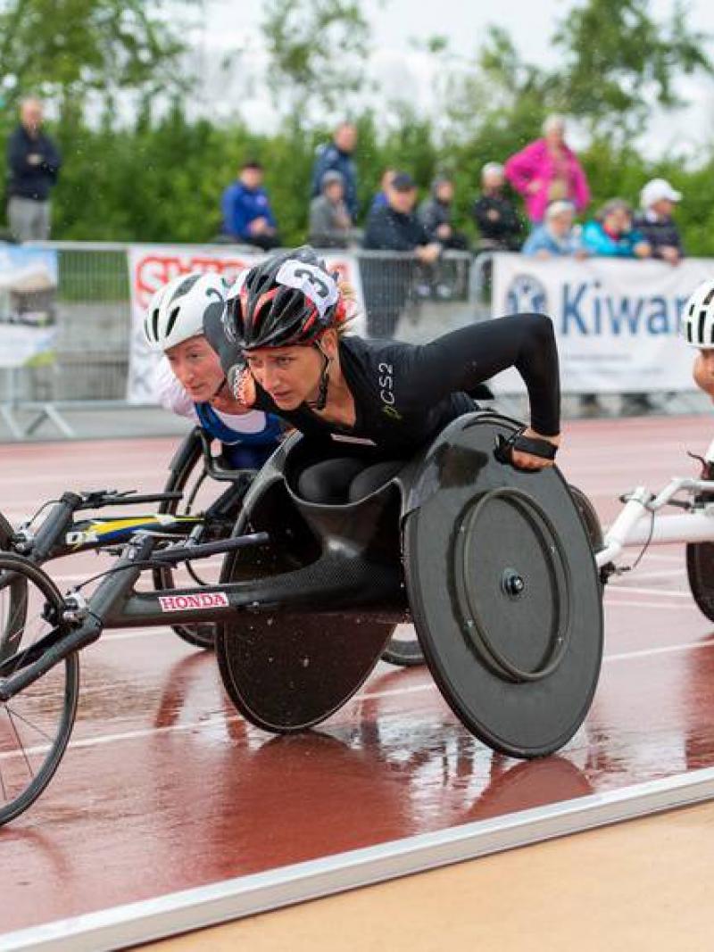 Three female wheelchair racers competing on a track under rain