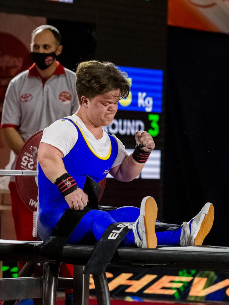 A female powerlifter celebrating seated on the bench press with two men standing behind her