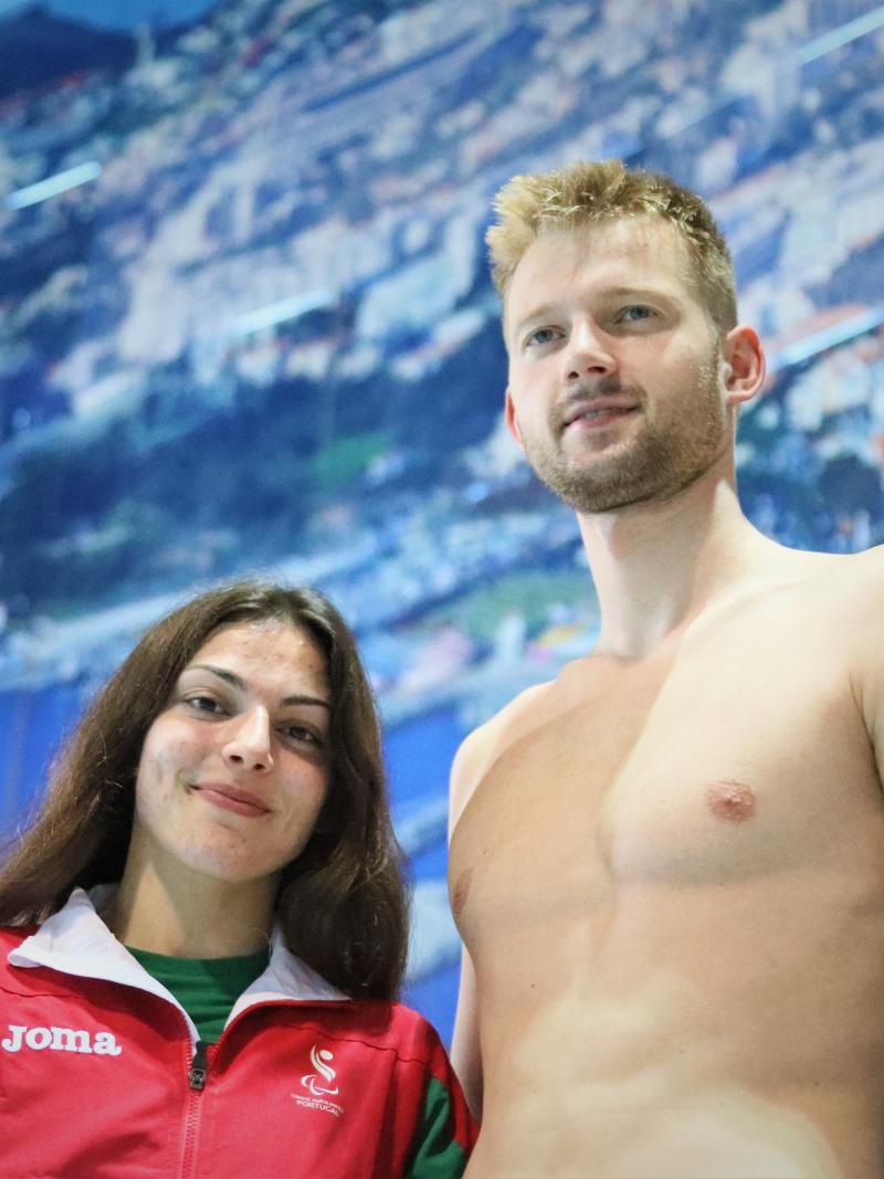 A short woman with a Portugal jumper and a tall shirtless man side by side in front of a panel in a swimming pool