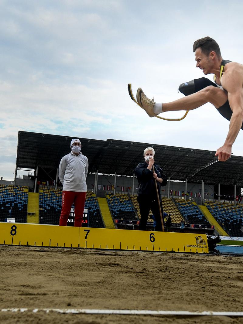 A men with a prosthetic leg jumping in a long jump event observed by three man