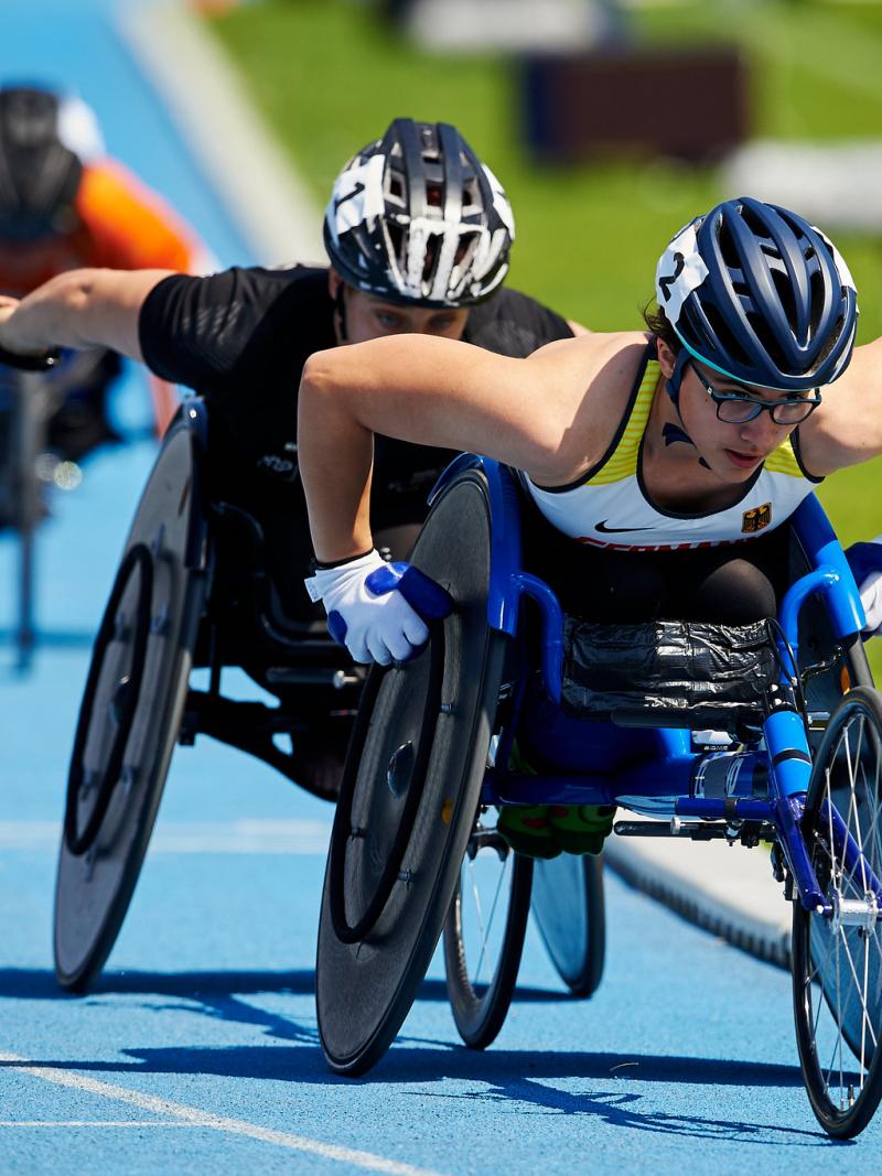 A woman in a racing wheelchair being followed by two athletes during a competition