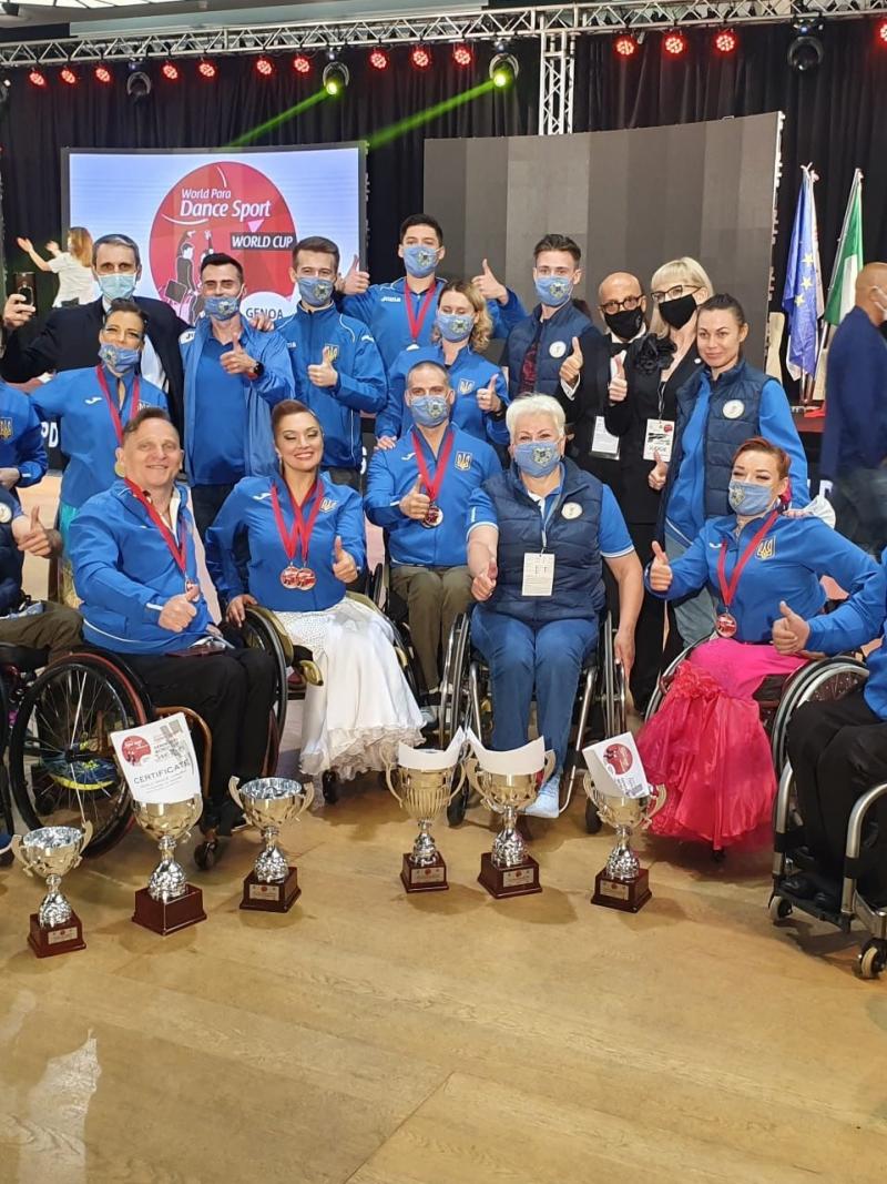 A group of nine people in wheelchairs with 12 people standing behind them posing for a picture with trophies in front of them