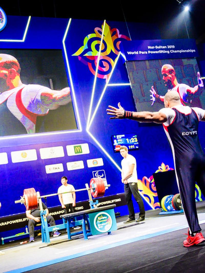A man standing in front of a bench press on a stage observed by six men standing and three persons behind a table
