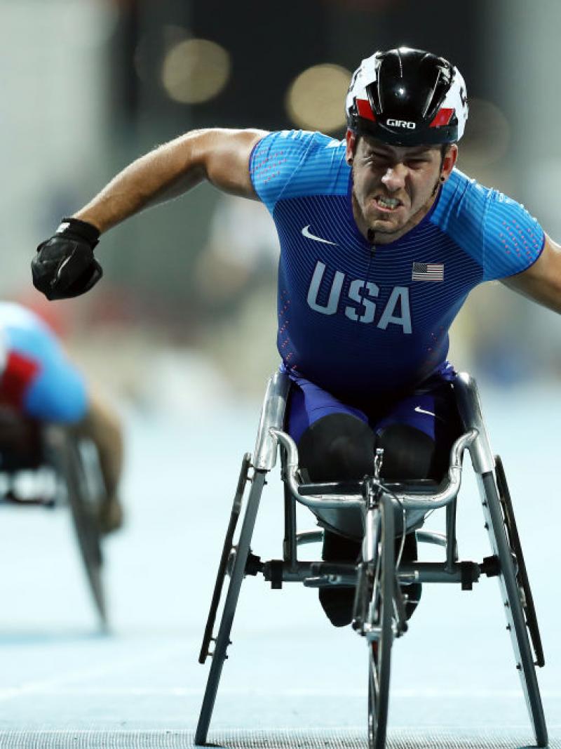 A man in a racing wheelchair celebrating with another competitor in the background
