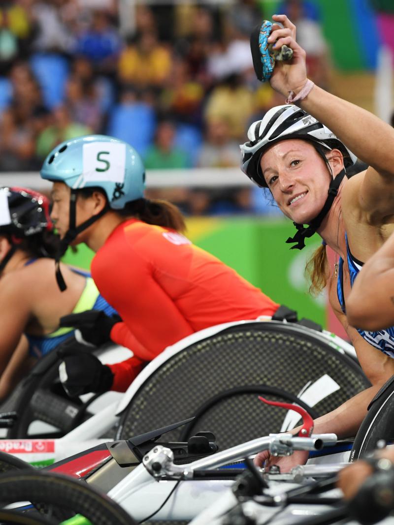 A woman in a racing wheelchair waving to the camera surrounded by other six female wheelchair racers