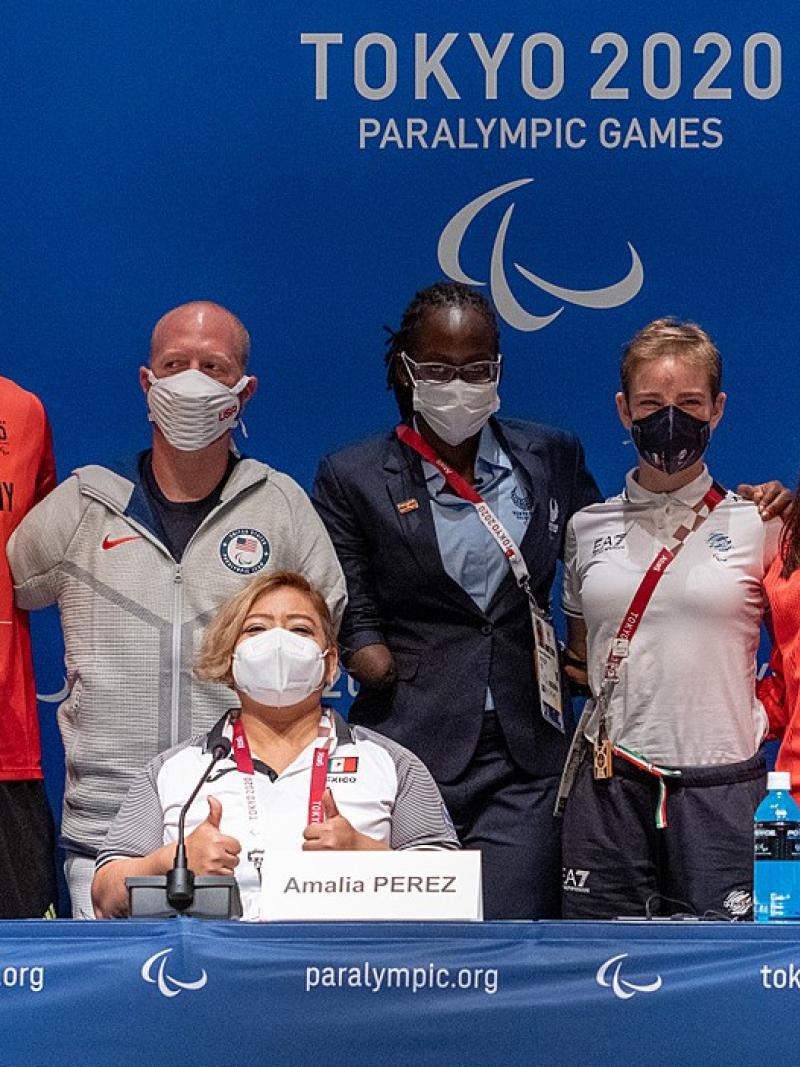 Two men and three women standing behind a seated woman in a press conference room