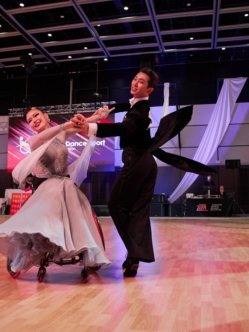 A female wheelchair dancer and her standing male partner in a Para dance sport competition observed by seven people standing in the background