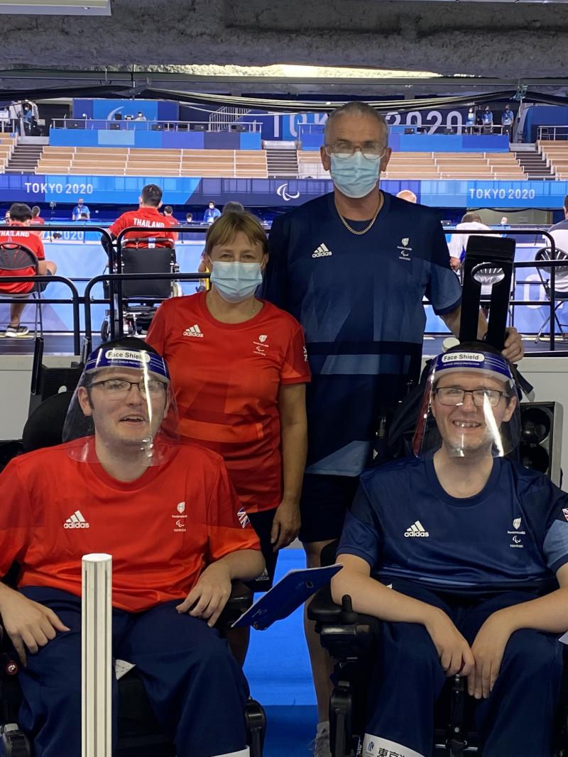 Linda stands behind Scott in his wheelchair, both wearing red, while Gary stands behind Jamie in his wheelchair, both in blue