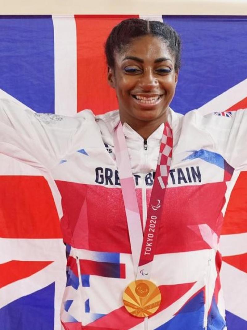 Kadeena Cox smiles with the gold medal hanging around her neck while holding the British flag