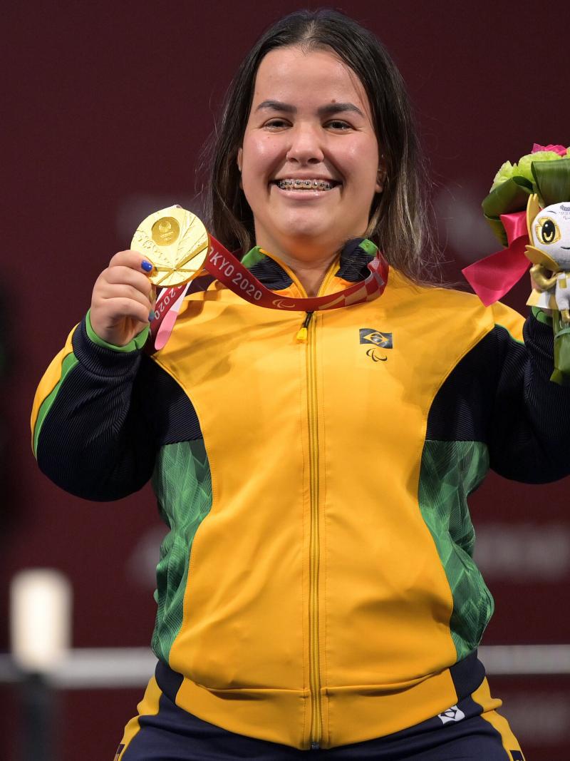 A woman posing with the gold medal in her right hand and a mascot in her left hand. She is having a big smile on her face