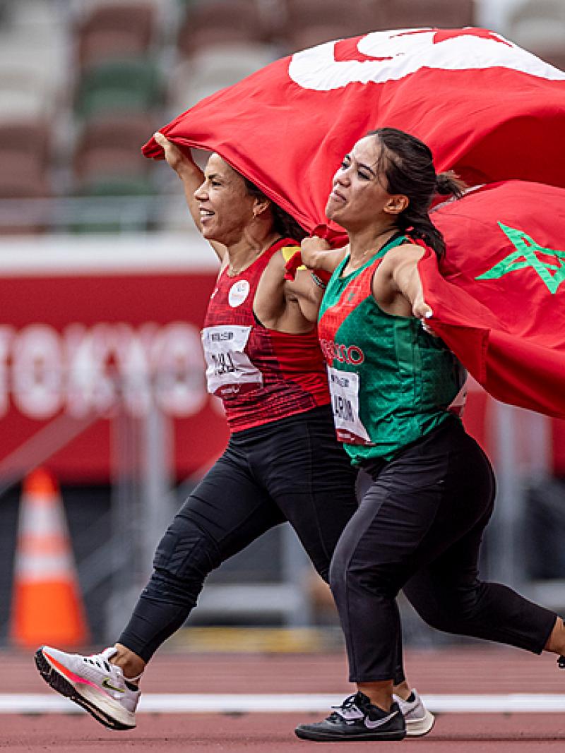 Two short stature women running on an athletics track holding the flags of Tunisia and Morocco