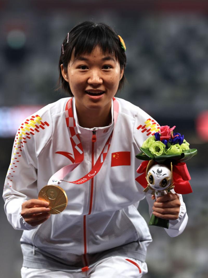 Xiaoyan Wen crouches on the podium displaying gold medal and mascot