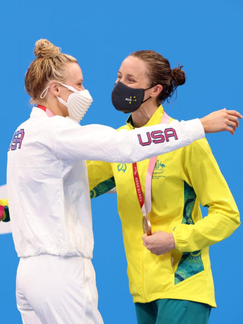Two women hugging in front of a backdrop showing the Paralympic Agitos in white