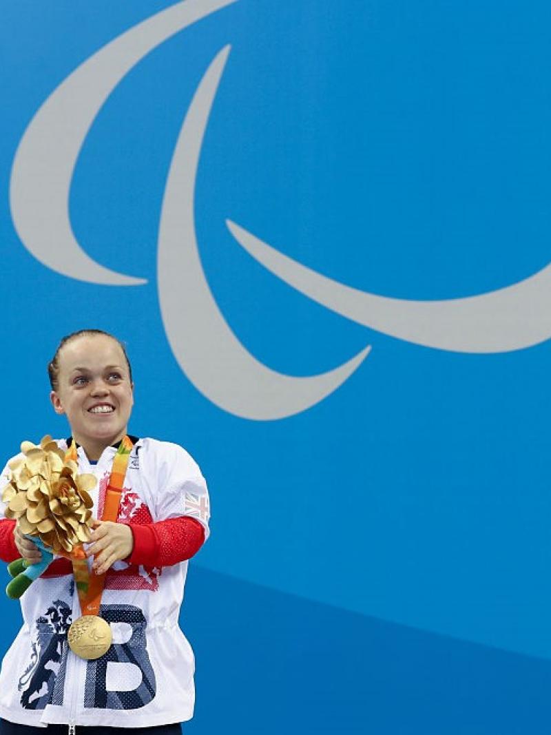 A short stature woman with her gold medal in front of a wall with the Paralympic Agitos