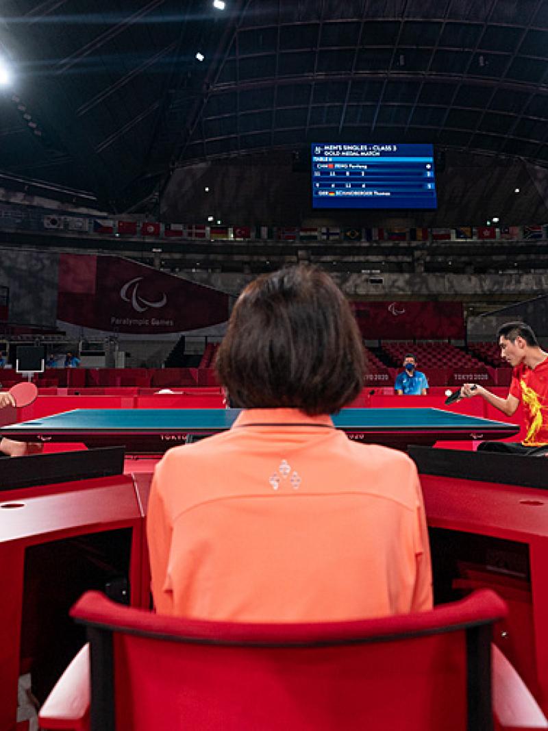 Two male wheelchair table tennis players playing with judge looking on