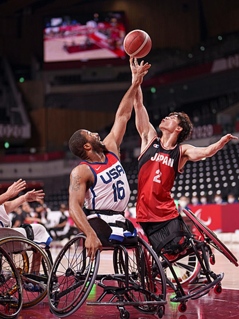 Two wheelchair basketball male athletes reach for the ball in the air