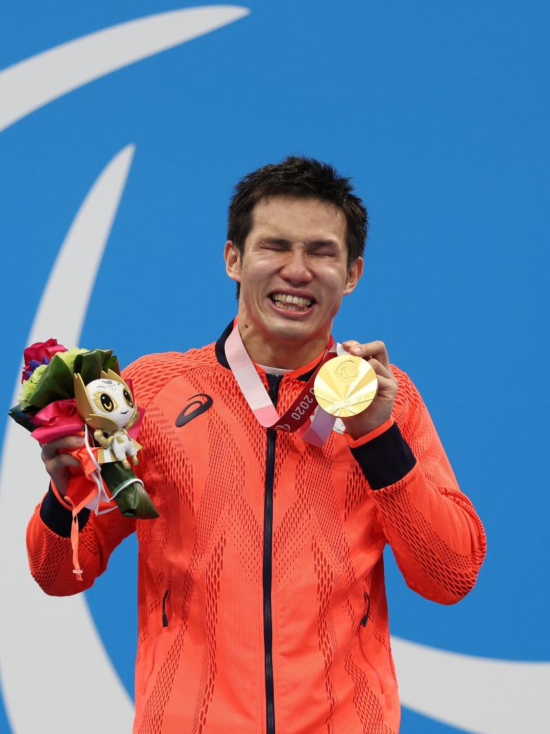 A vision-impaired athlete posing with a gold medal in left hand a mascot in his right hand