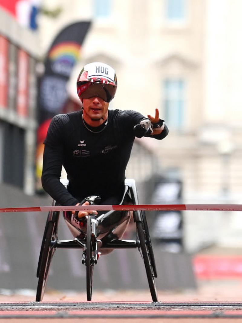 A man crosses the finish line in a wheelchair holding his left hand up with two fingers up signalizing victory