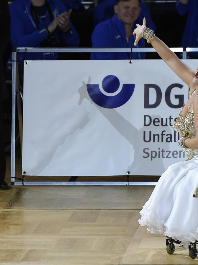 A woman in a white and gold dress dancing in a wheelchair with right arm raised