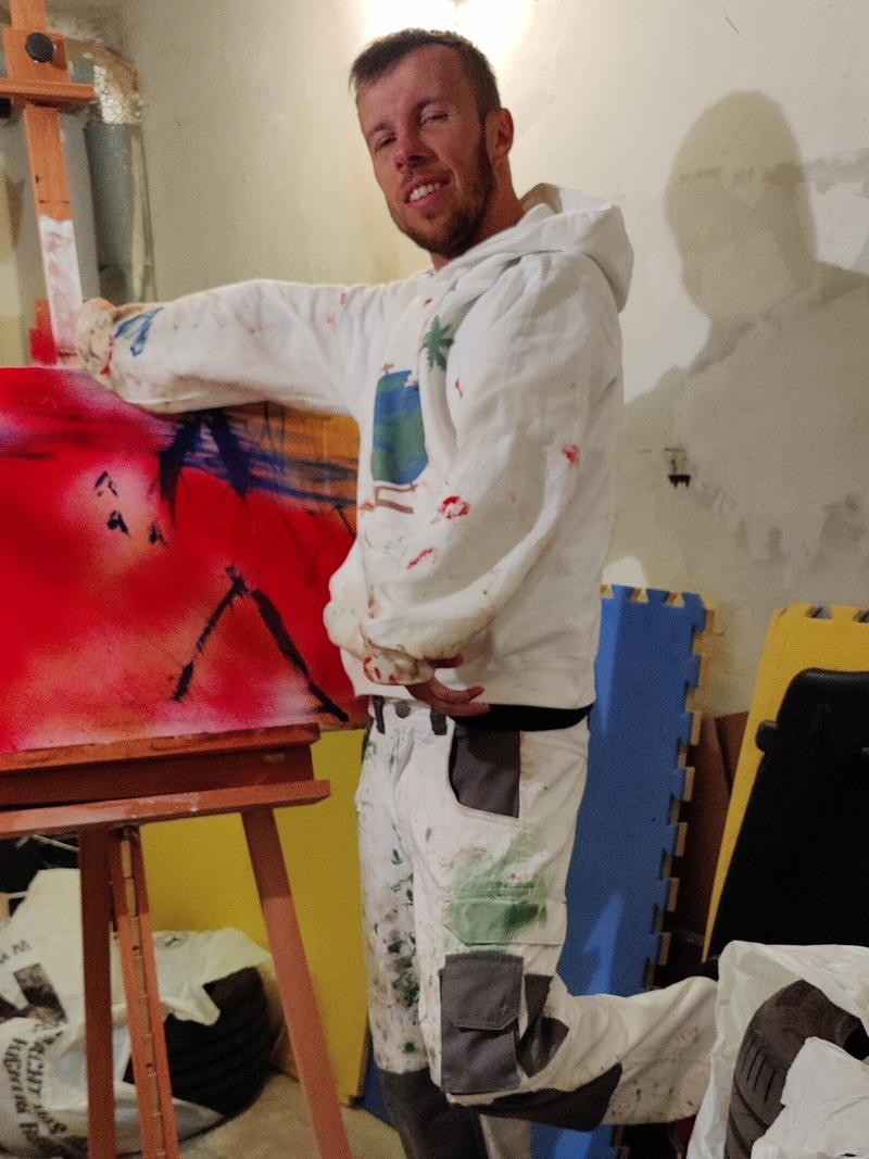 A man posing for a photo in front of his painting in a messy basement