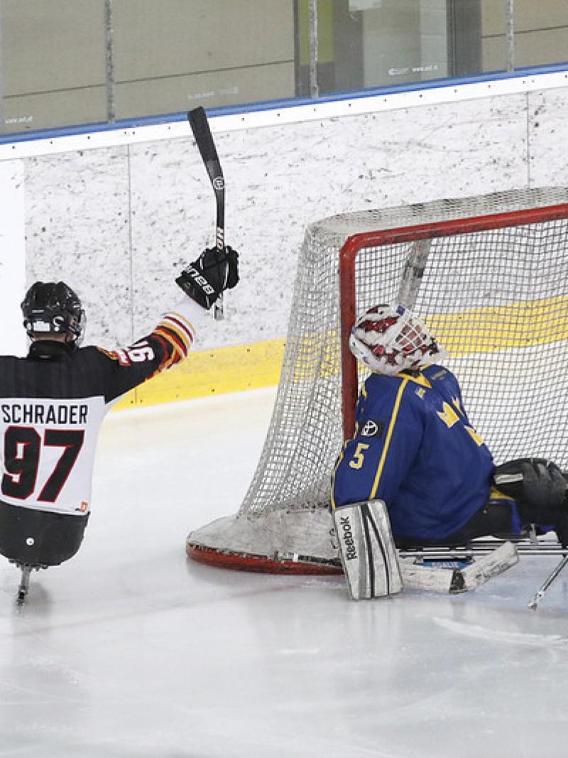 A German Para ice hockey player celebrating against two Swedish players on an ice rink