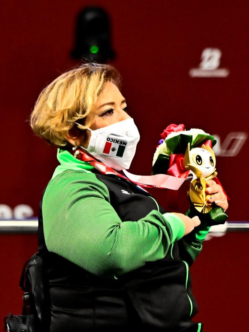 A woman in a wheelchair listening to the national anthem and holding a gold medal and a mascot in her left hand and holding her left hand high.