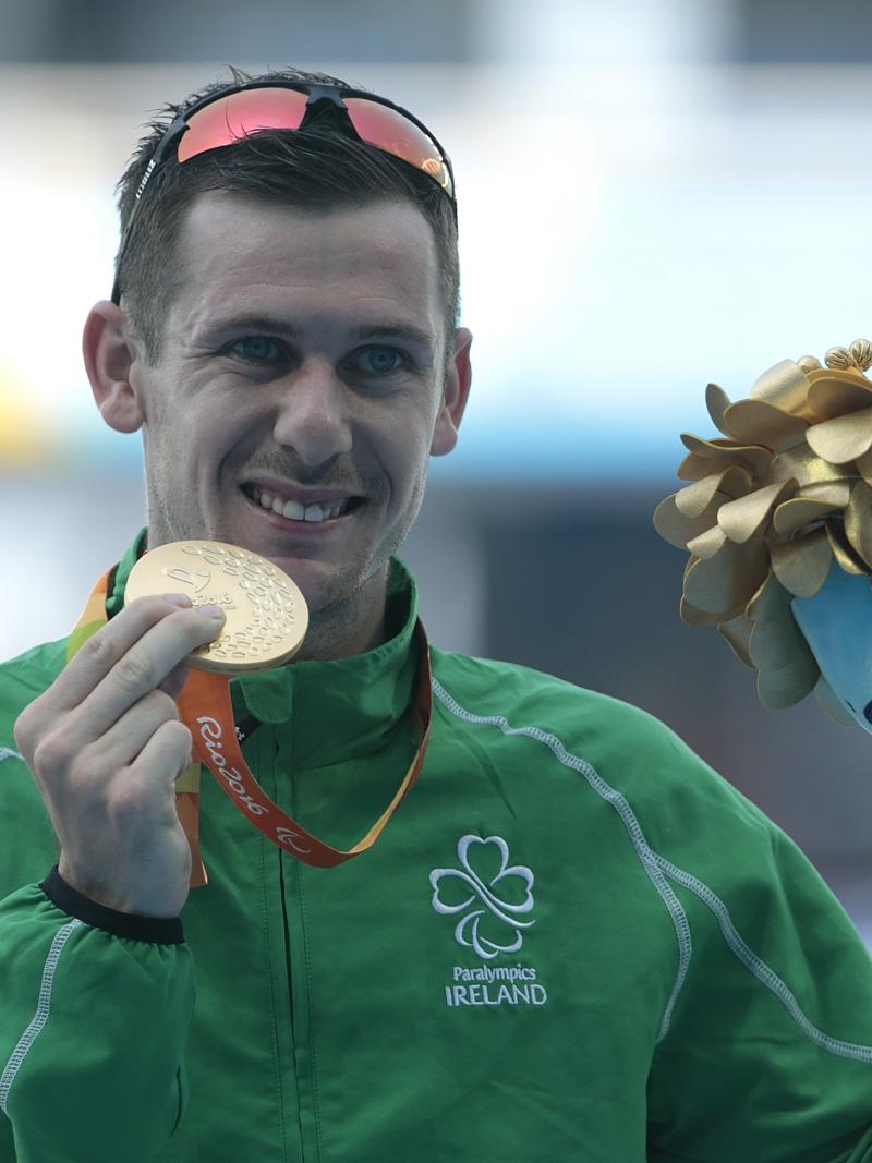 A man with the uniform of Ireland showing his gold medal and a mascot