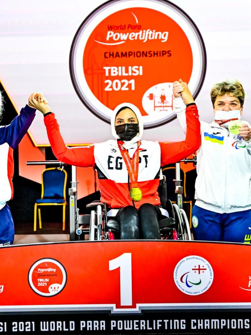 A woman in a wheelchair racing holding the arms of the short-stature women on a podium