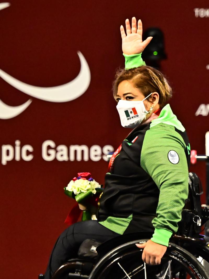 A woman in a wheelchair, raising her right hand high in celebration on the podium.