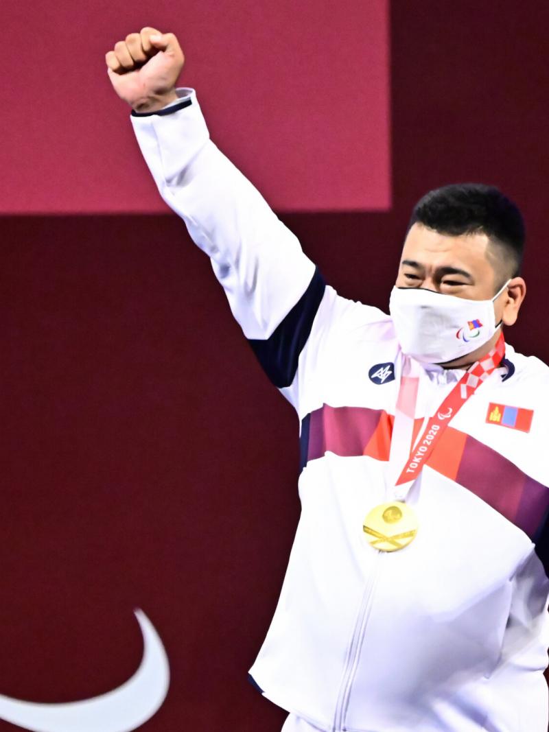 A man holding his right hand high with a gold medal around his neck and a mask on his face.