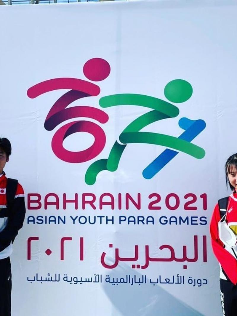 Two young Japanese Para athletes waving in front of a poster of the Asian Youth Para Games in Bahrain