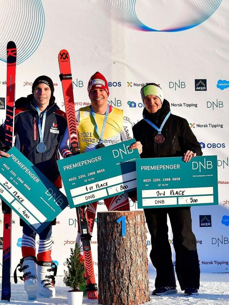 Three athletes standing in the snow in front of a white banner holding money prize.