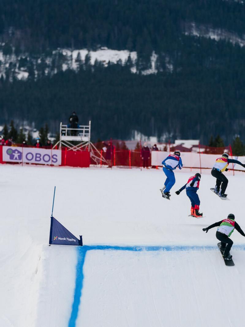 Four male athletes competing in a Para snowboard cross race. Three of them are jumping in the air, while the last one is about to jump.