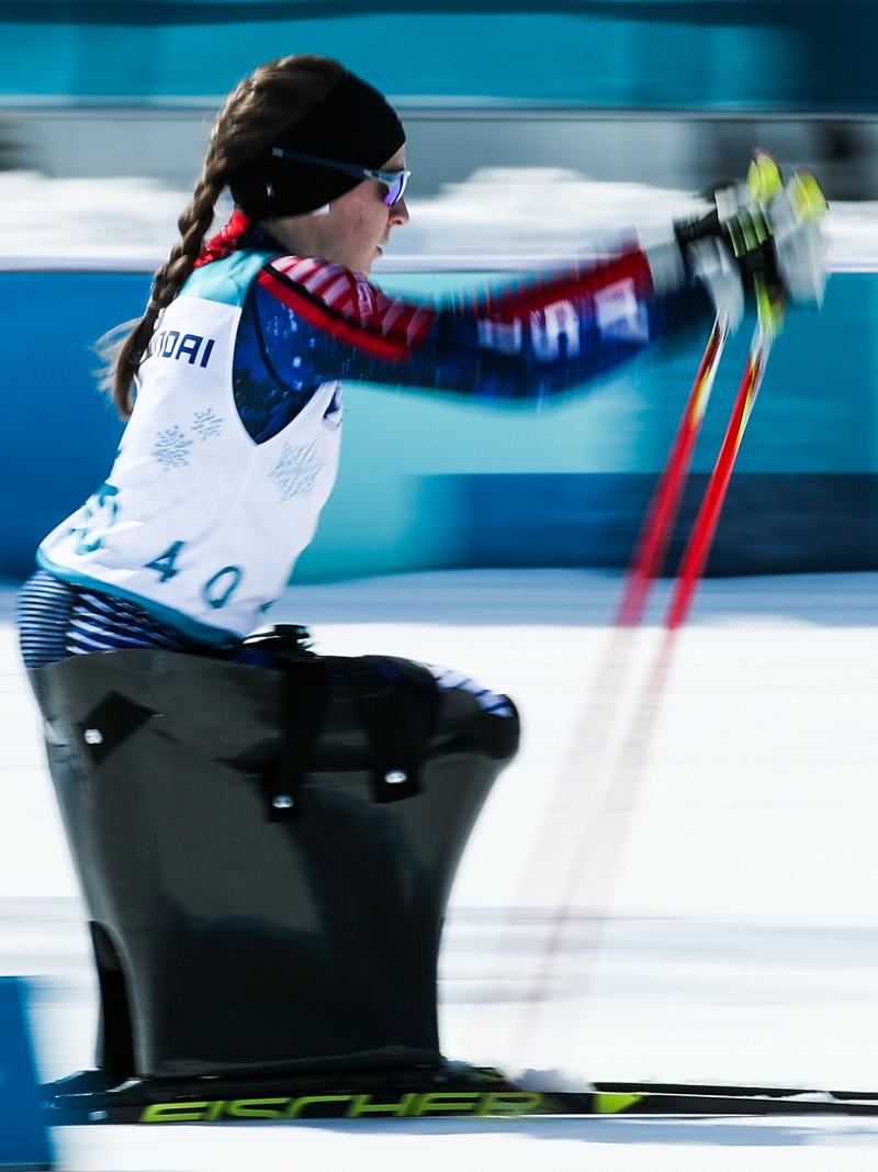 A woman in a sit ski competition pushing herself forward with two sticks.