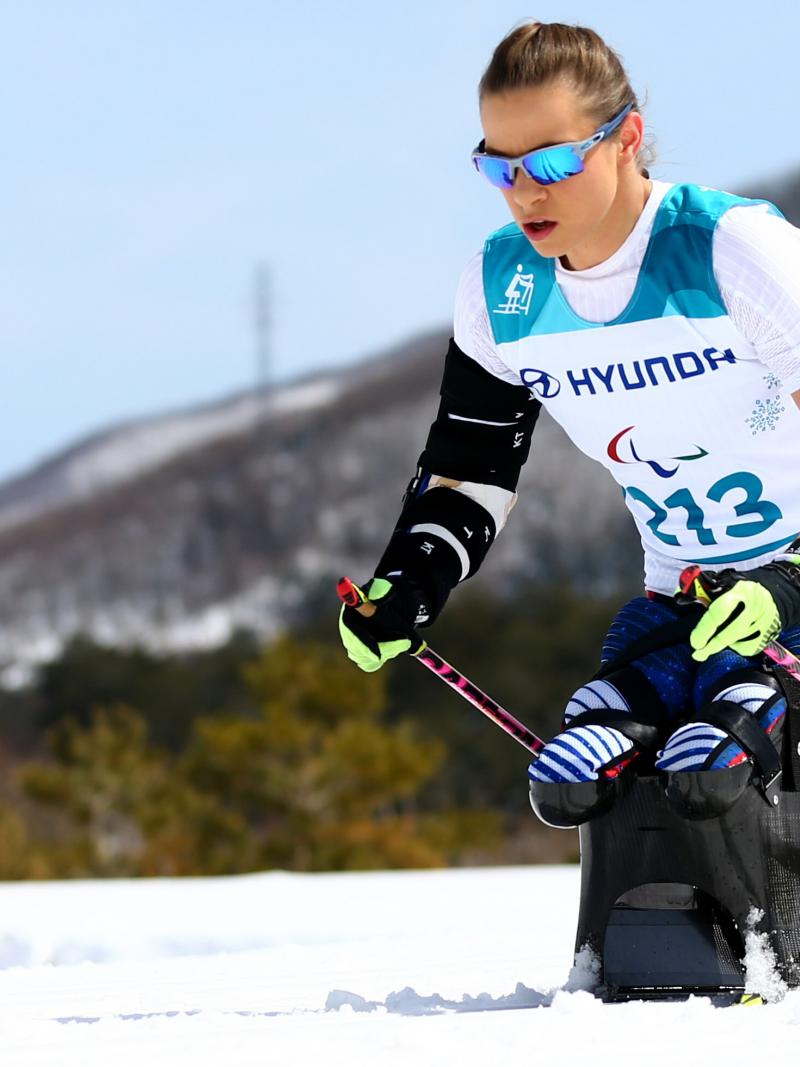 A woman with goggles pushing herself forward with two sticks in a sit ski competition on a sunny day.