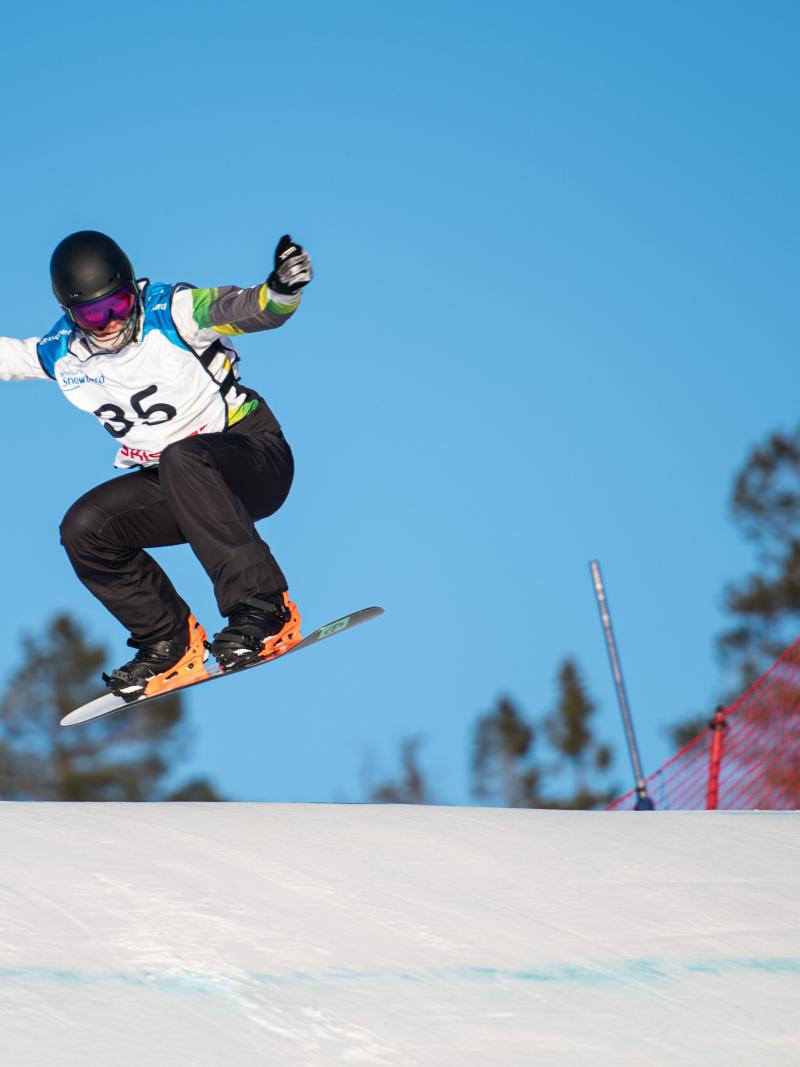 A man on a snowboard jumping into the air with his arms raised on a sunny day in the snow.
