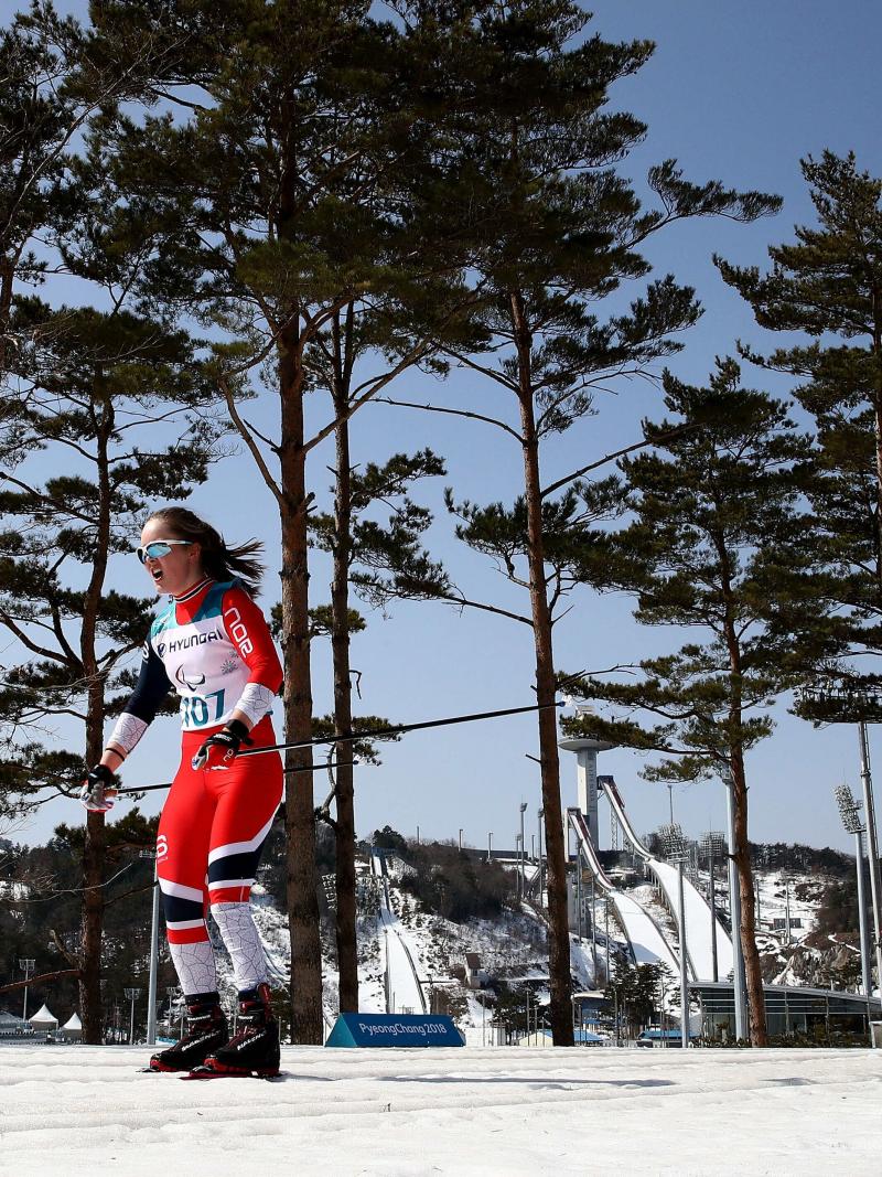 A woman in a cross-country race with goggles on her head during a sunny day. Trees are in the background with the clear and blue sky.