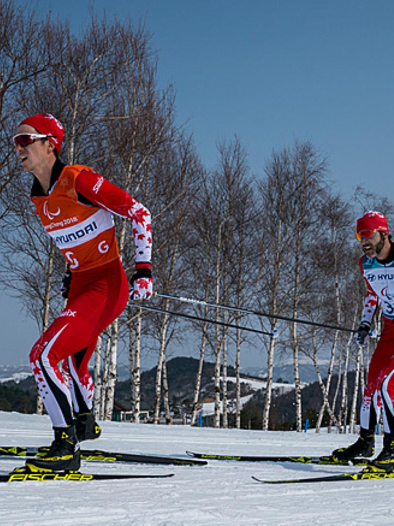 A male cross-country skier behind his male guide in a Para cross-country skiing race
