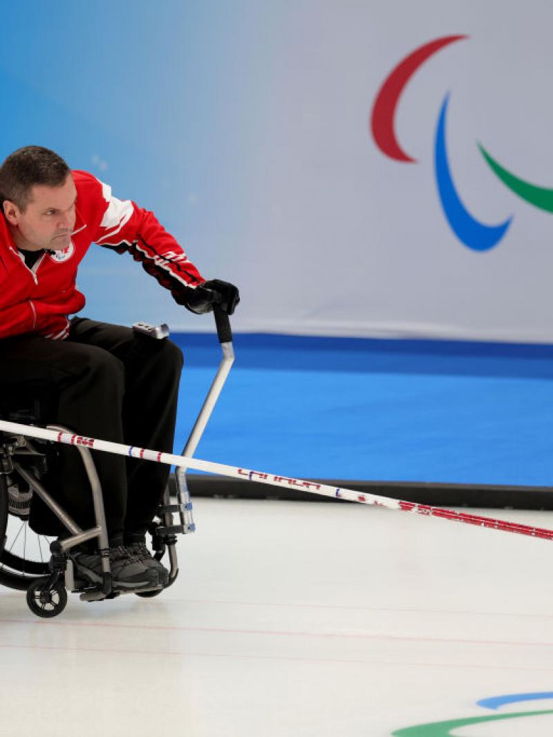 Mark ideson of Canada is held by a teammate as he goes to release the curling stone at Beijing 2022