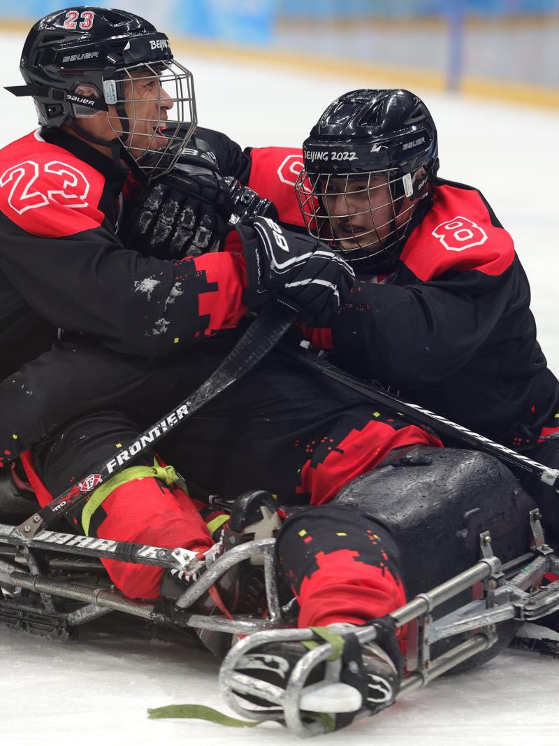 Three men on sleds lying on the ice and hugging each other in full Para ice hockey gear.