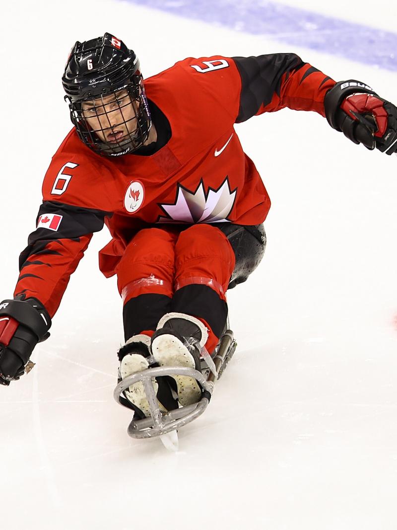 A male player on a sledge kicks the puck with a stick.