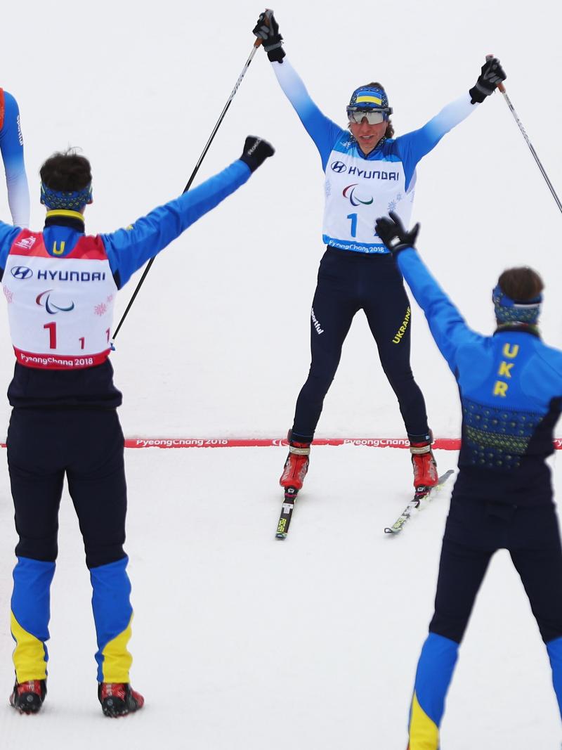 A woman crosses the finish line with raised hands and two sticks in them as the team cheers her on.