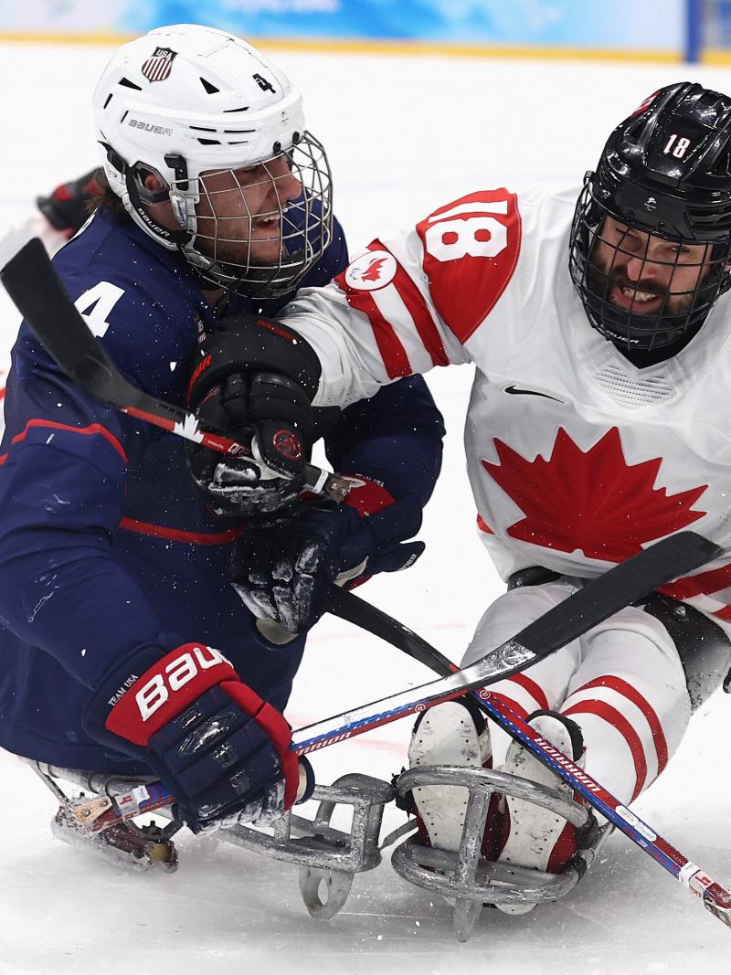 Two Para ice hockey players in full gear, one in white and the other in the blue jersey, pushing each other to gain possession of the puck.