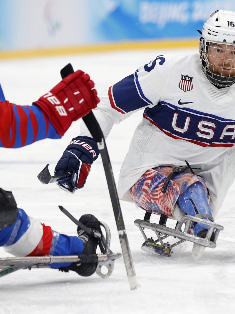 A Para ice hockey player in white and blue jersey hits the puck with a stick in left hand.