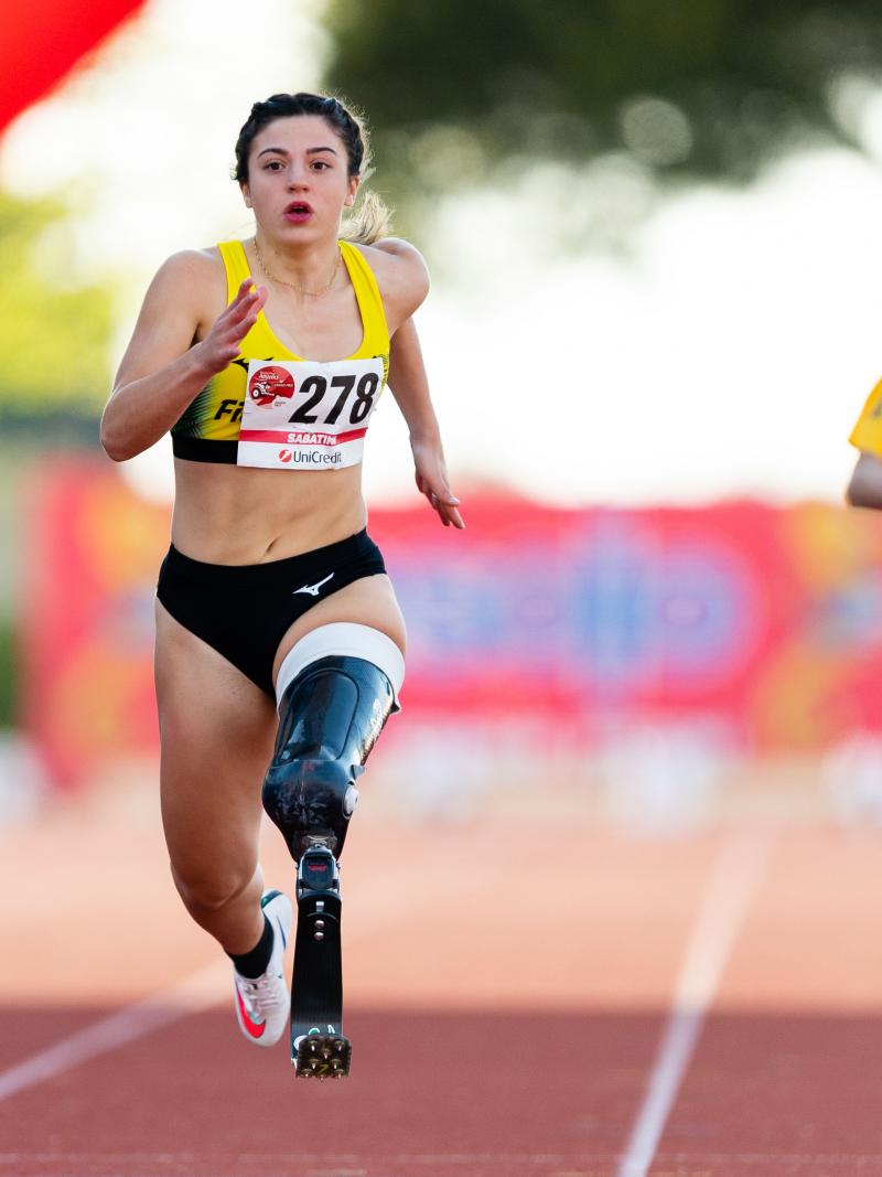 A female athlete with her left leg amputated above the knee running on the track in front of two other female athletes.