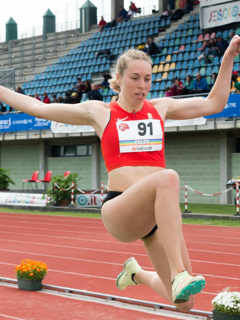 A woman in a red top jumping at the stadium with both of her hands in the air.