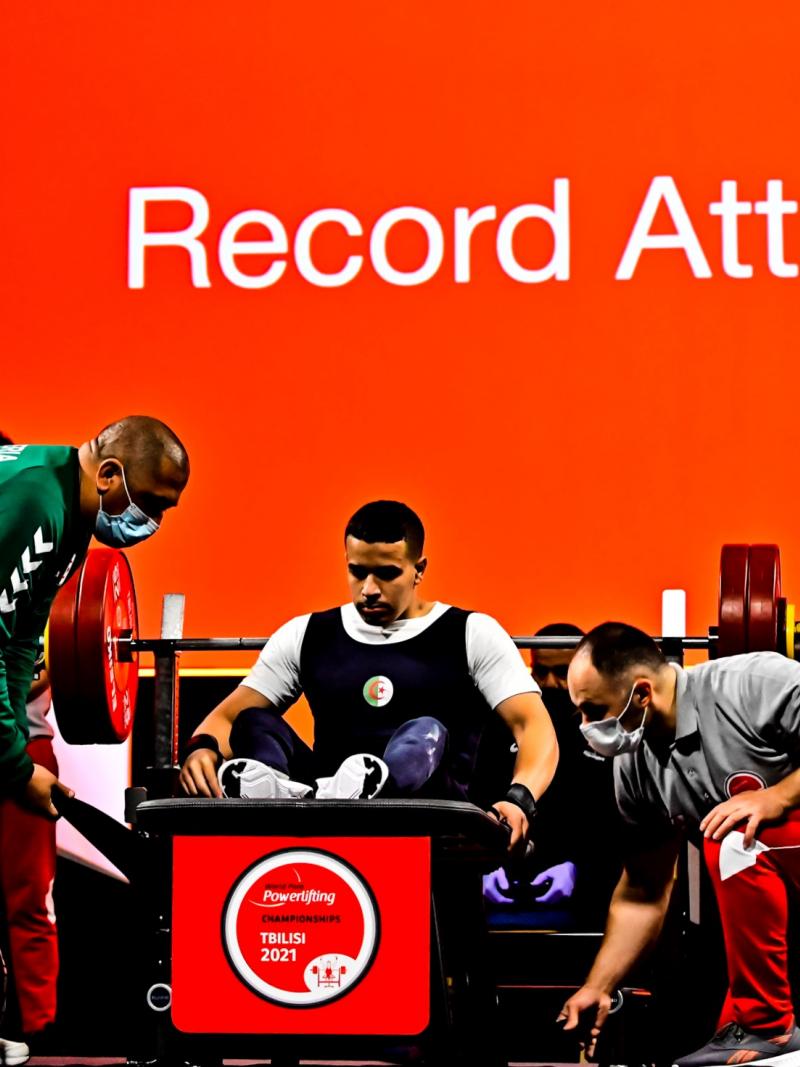 A Para powerlifter sitting on the bench with two other men wearing masks and tying the legs of the athlete sitting on the bench.