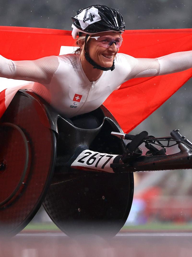 A woman in a racing wheelchair holding the Swiss flag with a smile on her face.