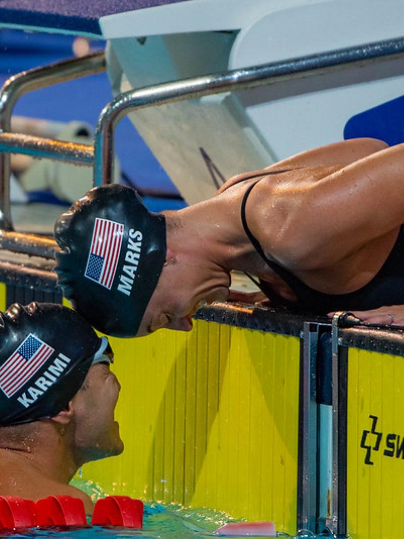 A woman outside a swimming pool stretching her head to touch the head of a man inside the pool