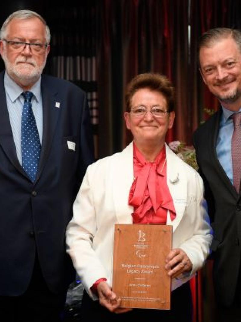 Two men in suits stand on both sides of a woman holding an awards plaque.